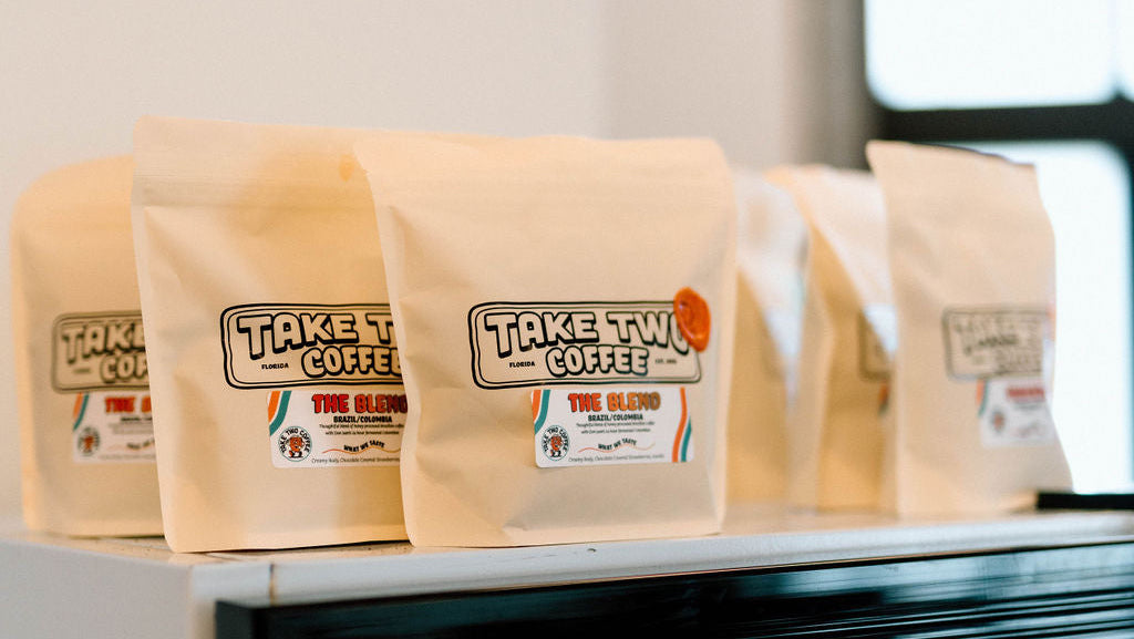 Packages of Take Two Coffee on a shelf with a refrigerator displaying more coffee products in the background.
